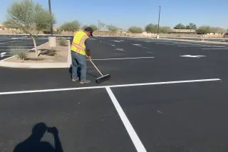 Worker sweeping a freshly paved asphalt parking lot with white stall lines under the sun in Goodyear, AZ.