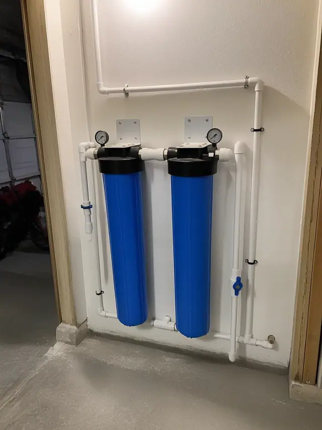 Whole house carbon water filtration system with blue filter housings installed on a garage wall in Mesa, AZ.