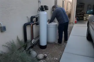 Technician inspecting copper plumbing and a water softener unit beside a garage in Mesa, AZ.