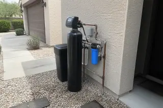 Water softener unit with plumbing hardware installed against a stucco wall and desert landscaping in Mesa, AZ.