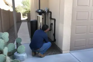Technician checking saltless water softener plumbing near a garage and cactus in Mesa, AZ.