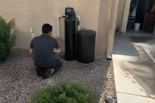 Technician inspecting water softener pipes and valves beside a garage in Mesa, AZ.