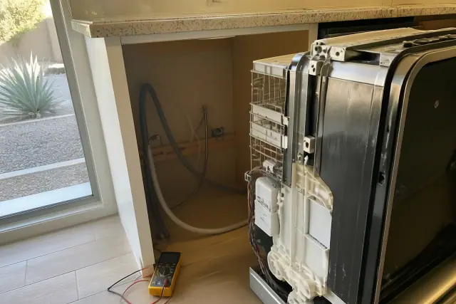 Technician repairing a dishwasher in a kitchen in Chandler, AZ, with exposed hoses and a multimeter on the floor.