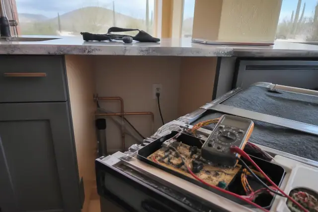 Technician repairing a dishwasher with exposed wiring and plumbing in a kitchen in Goodyear, AZ.