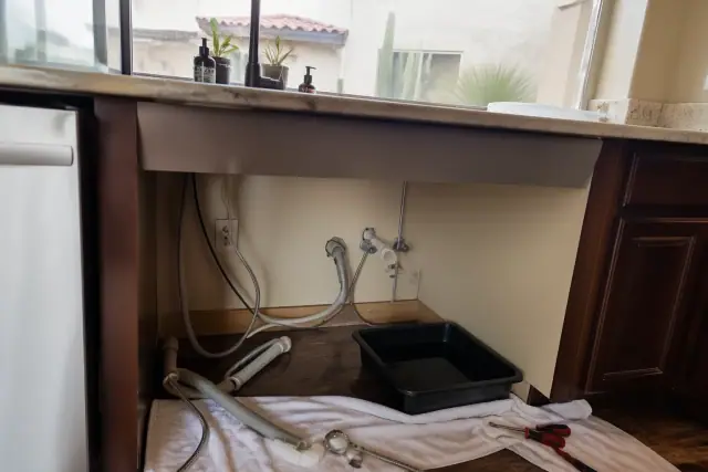 Repairing a dishwasher drain clog and pump in a Phoenix, AZ kitchen with exposed plumbing and a catch pan on the floor.