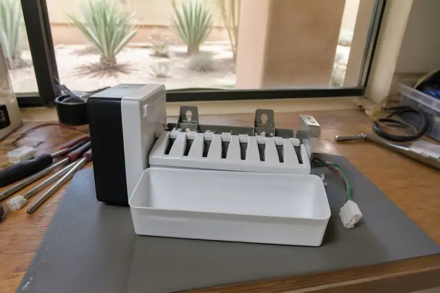 A technician repairing a freezer ice maker on a workbench in Phoenix, AZ, with desert landscaping visible through a window.