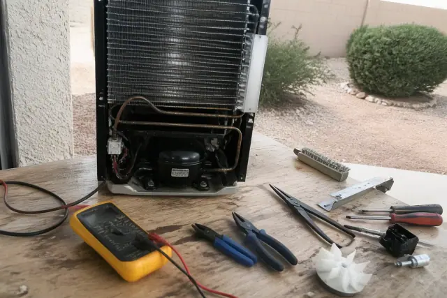 Technician repairing a disassembled freezer cooling unit with tools and a multimeter at a workbench in Avondale, AZ.