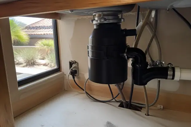 Garbage disposal repair under a kitchen sink in Goodyear, AZ with desert landscaping visible through the window.