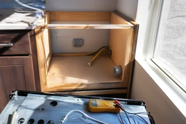 Technician repairing a gas oven in a wooden cabinet cutout with a multimeter and gas line visible in Phoenix, AZ.