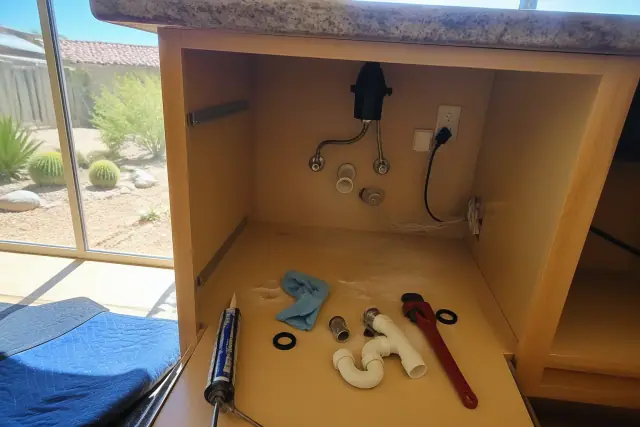 Disassembled P-trap, basin wrench, and sealant under a kitchen sink during a repair in Phoenix, AZ.