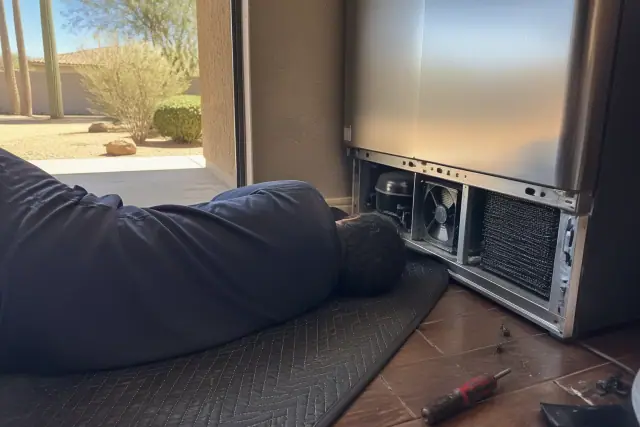 Technician inspecting a refrigerator condenser and maintenance brackets in a Phoenix, AZ home.
