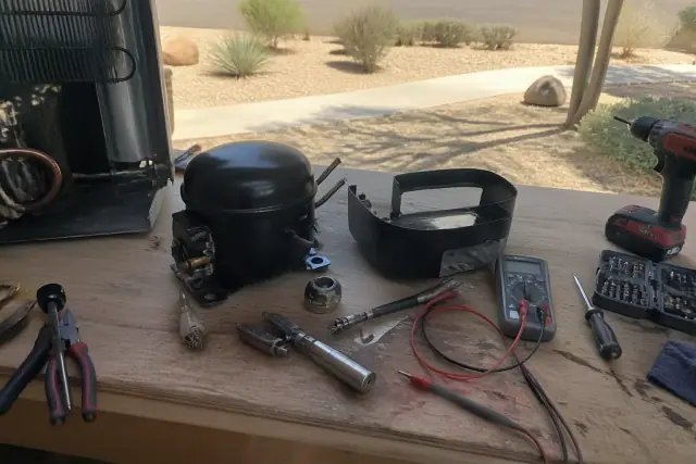 Disassembled refrigerator compressor and multimeter on a workbench during a repair in Paradise Valley, AZ.