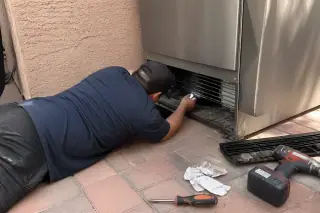 Technician inspecting the condenser coils at the base of a stainless steel refrigerator in a Phoenix, AZ home.