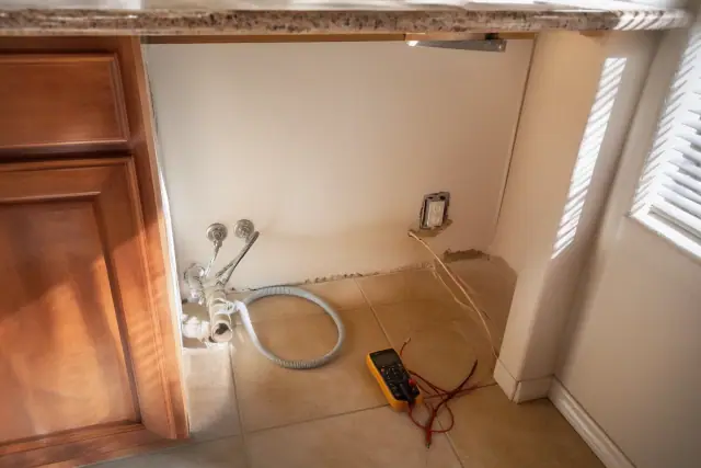 Empty kitchen dishwasher space with exposed plumbing, wiring, and a multimeter on the floor in a Phoenix, AZ home.