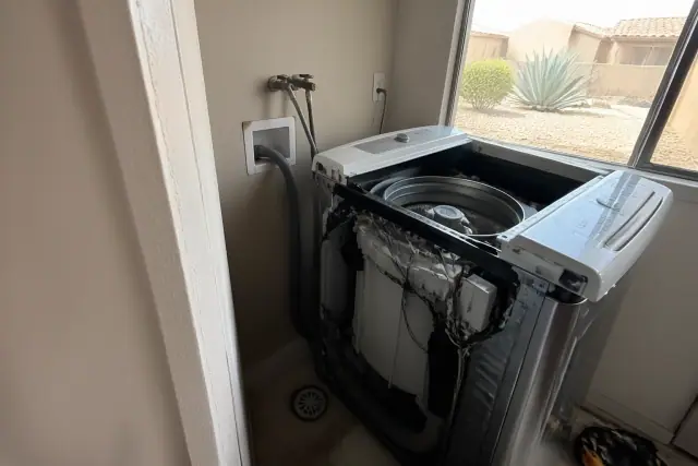 A technician repairing a washing machine in a Glendale, AZ utility alcove with desert landscaping visible outside.