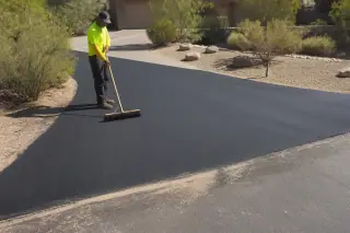 Worker sweeping a newly paved asphalt driveway transition in Phoenix, AZ, under bright sunlight.