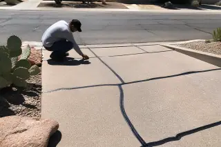 Technician inspecting crack sealant on a sloped driveway transition near prickly pear cacti in Phoenix, AZ.