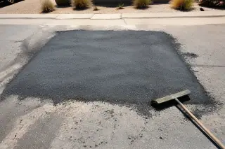Newly compacted asphalt utility patch with loose aggregate and a broom in Phoenix, AZ, near desert gravel landscaping.