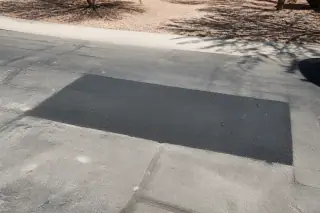 Rectangular asphalt utility patch in a city road in Phoenix, AZ, surrounded by sun-bleached desert gravel.