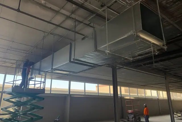 Sheet metal workers installing galvanized steel HVAC ductwork from a high ceiling in a Peoria, AZ industrial warehouse.