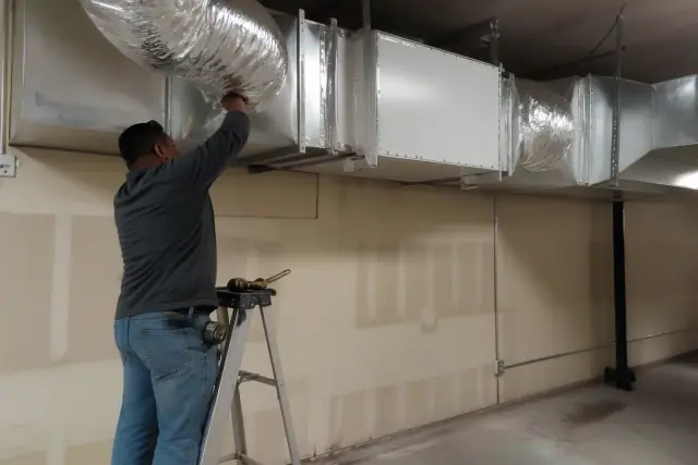 Installer securing a commercial air purification unit to ventilation ductwork in a Phoenix, AZ warehouse.