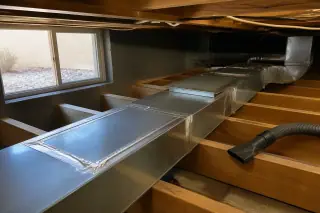 Clean silver metal air ducts and a vacuum hose nozzle on wooden joists in a Phoenix, AZ basement.