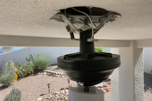 Electrician installing a ceiling fan bracket and wiring in a Tempe, AZ home with desert landscaping visible outside.