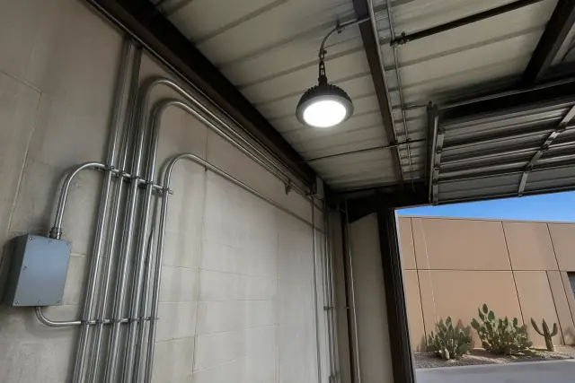 Newly installed industrial LED high-bay light and conduit pipes on a stucco wall at a workspace in Phoenix, AZ.