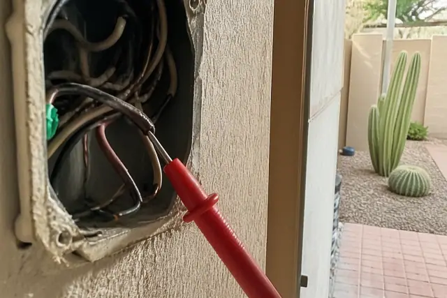 Multimeter probe testing damaged wires in a Scottsdale, AZ residential utility closet with desert landscaping outside.