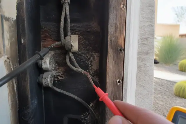 Multimeter probe testing frayed, charred knob-and-tube wiring inside a dark wall in a Phoenix, AZ home.