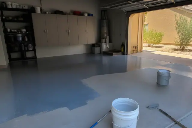 Contractor applying epoxy to a residential garage floor in Phoenix, AZ, with utility shelving and buckets in view.