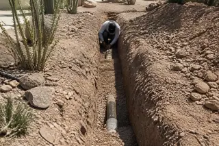 Worker inspecting a straight utility trench with exposed pipes in rocky soil at a Phoenix, AZ construction site.