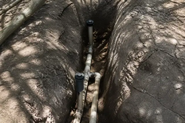 Exposed PVC irrigation pipes and sprinkler heads in a narrow soil trench under a palo verde tree in Phoenix, AZ.