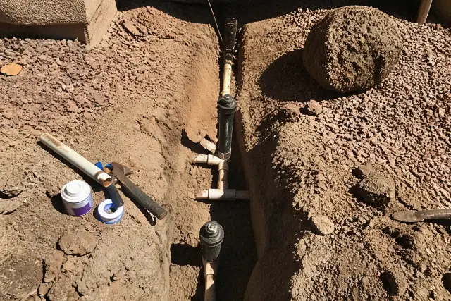 Excavated trench showing irrigation pipe repairs and hand tools in a residential yard in Phoenix, AZ.