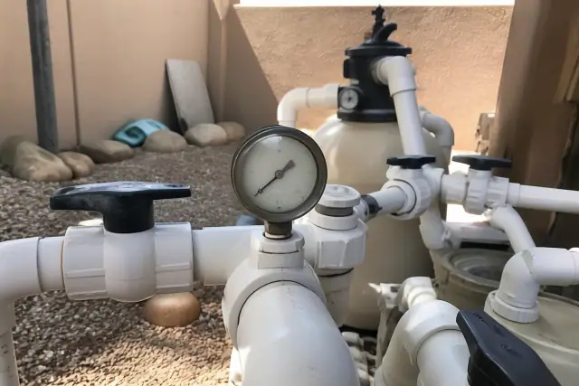 Pool plumbing manifold with a pressure gauge and PVC pipes in a Phoenix, AZ backyard with xeriscaping.