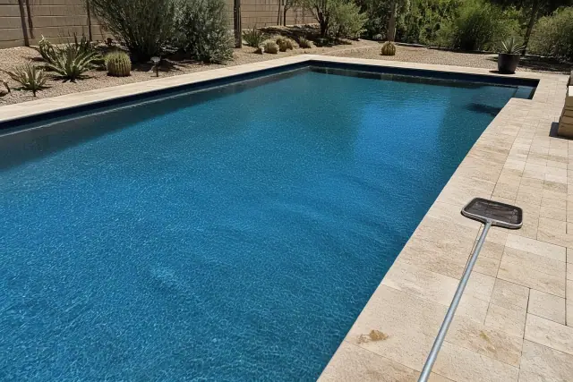 A crystal clear backyard pool in Phoenix, AZ, after professional debris cleaning with a net resting on travertine pavers.