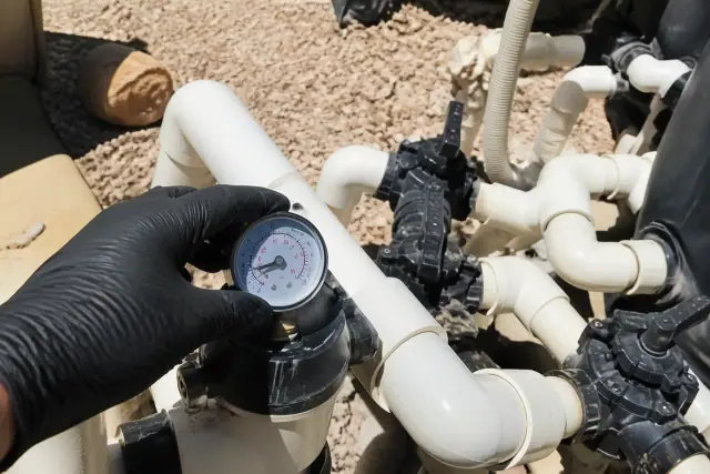 Technician's gloved hand checking a pool pressure gauge on a PVC manifold during service in Phoenix, AZ.
