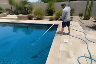 Technician using a net to clean leaves from a rectangular pool in a Phoenix, AZ backyard with desert landscaping.
