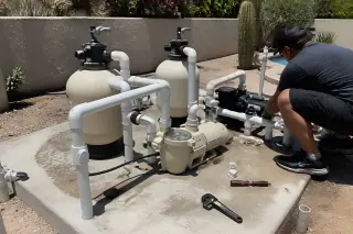 Contractor inspecting repaired pool pump equipment and PVC plumbing on a concrete slab in a Phoenix, AZ backyard.
