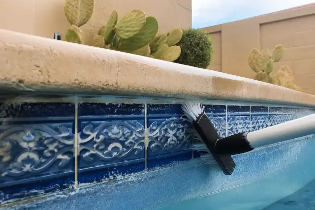 A technician scrubbing mineral deposits from pool waterline tiles at a home in Glendale, AZ with desert landscaping nearby.
