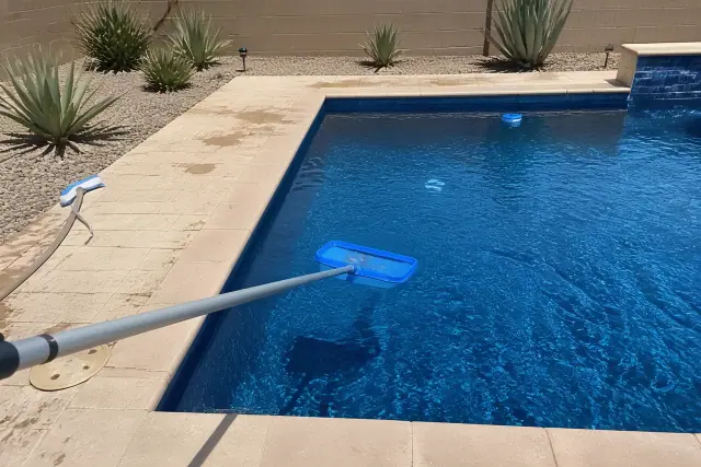 A telescoping net pole skimming a rectangular pool on a tan paver deck in Phoenix, AZ.