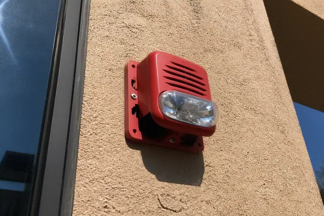 A wall-mounted fire alarm horn-strobe device being installed on a stucco exterior wall in Phoenix, AZ.