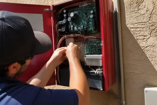 Technician repairing wiring inside a commercial fire alarm control panel on a stucco wall in Phoenix, AZ.