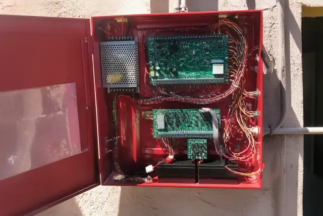 Open fire alarm control panel cabinet with multicolored wiring and circuit boards on a stucco wall in Mesa, AZ.