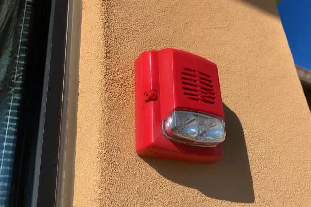 Wall-mounted fire alarm horn-strobe device installed on a stucco exterior wall in bright sunlight in Phoenix, AZ.
