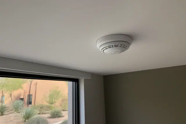 Dual-sensor smoke and CO alarm mounted on a ceiling in a Phoenix, AZ building with desert views through the window.