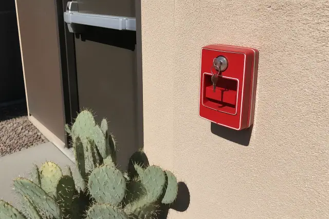 Red fire alarm pull station with a test key inserted on a stucco wall in Mesa, AZ, framed by prickly pear cactus.