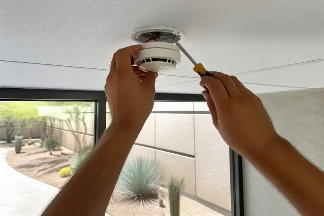 Technician mounting a smoke detector onto a commercial ceiling in Glendale, AZ with desert landscaping in the background.