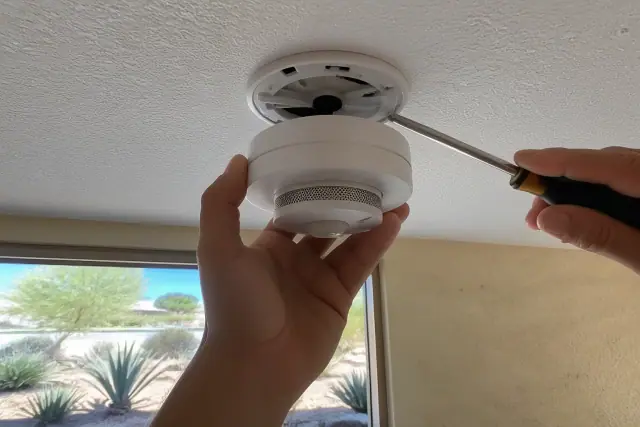 A smoke detector being installed on a commercial ceiling in Glendale, AZ with desert landscaping visible in the background.