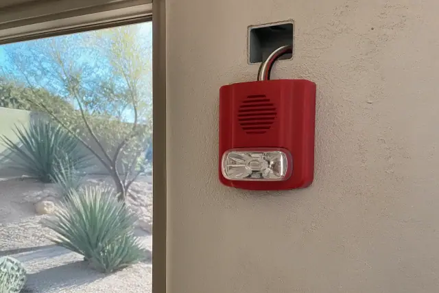 Wall-mounted fire alarm horn-strobe device being installed on a stucco exterior wall in Peoria, AZ.